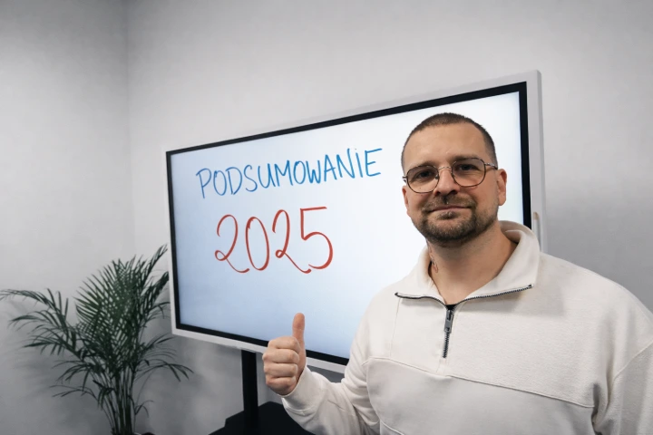 Mężczyzna w jasnej bluzie i okularach stoi w nowoczesnym biurze obok dużego ekranu z napisem „Podsumowanie 2025” i pokazuje kciuk w górę. W tle jasna ściana i roślina doniczkowa.